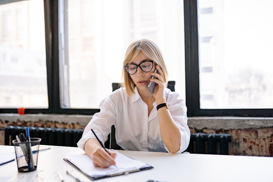 A thoughtful professional in an office setting making a phone call, with a visible notepad for jotting down voicemail notes.